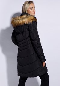 Black padded coat with a detachable tan fur hood, featuring vertical stitching, long sleeves, and a flared hemline.