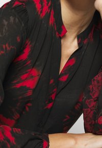 Kobieta w czarnej i czerwonej bluzie w wzór tie-dye z guzikami i kołnierzykiem mandarynkowym, opierająca ramię na powierzchni.