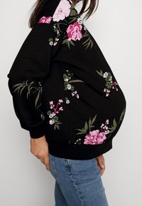 Mujer embarazada con un sudadera negra con patrones florales en rosa y blanco, combinada con jeans azules, mostrada en perfil lateral.