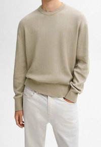 Mann in einem beigen gerippten Langarmpullover und hellen Hosen, mit einer Hand in der Tasche und neutralem Hintergrund.