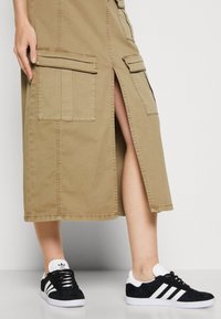 Onderbenen met zwarte en witte Adidas sneakers, gecombineerd met een beige middellange rok met grote voorzakken en een voorsplit.