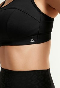 Fekete sportmelltartó sima anyagból, varrott varratokkal és egy kis fehér logóval az oldalon. Magas derékú leopárdmintás leggingsszel párosítva.