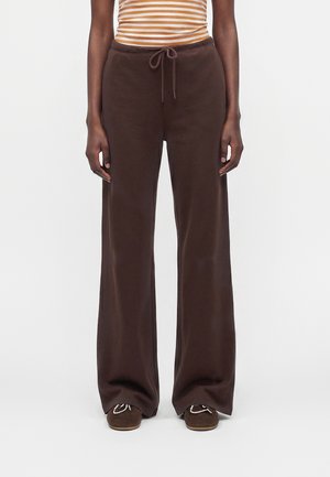 Person iført brune bukser med vide ben og snøre i taljen, stribet orange og hvid top og brune mokkasinslippers med syning.