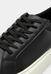 Gros plan d'une basket en cuir noir avec des lacets noirs et une semelle blanche, mettant en valeur les coutures détaillées et la texture.