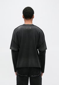 Fekete, túlméretezett pulóver réteges rövid ujjakkal és kopott szegéllyel, díszítő ezüst színű fémes elemekkel a nyakkivágás közelében.