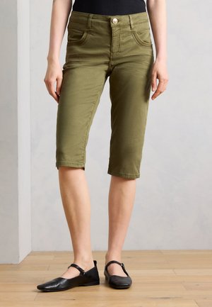 Kvinde iført olivengrønne knælange shorts og sorte Mary Jane-flade sko, stående på et lyst trægulv foran en ensfarvet væg.