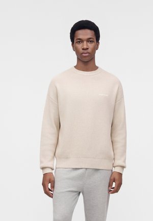 Un homme porte un pull en maille couleur crème et un pantalon gris clair sur un fond blanc uni.