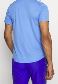 Camisa de manga corta azul claro en un tejido sintético suave, con un dobladillo redondeado y detalles en el yugo trasero, combinada con pantalones cortos deportivos morados.