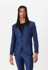 Costume bleu ajusté avec fermeture à deux boutons, cols crantés et poches latérales ; porté par-dessus une chemise noire. Texture lisse, coupe slim.