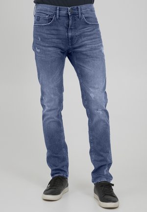 Schmal geschnittene blaue Jeans mit leichtem Used-Look, kombiniert mit schwarzen Turnschuhen vor einem schlichten grauen Hintergrund.