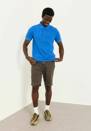 Junger Mann lächelt und richtet sein hellblaues Polohemd, trägt braune Shorts, weiße Socken und olivgrüne Turnschuhe.