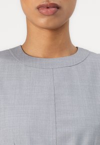 Grå, teksturert stoff med en glatt overflate. Har rund hals og detaljer med sentralsøm i front. Minimalistisk design.