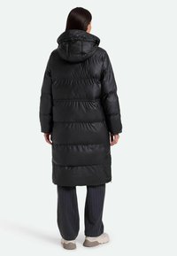 Cappotto imbottito nero con cappuccio, caratterizzato da un design trapuntato, una silhouette lunga e tasche con zip. Indossato sopra pantaloni larghi a fantasia.