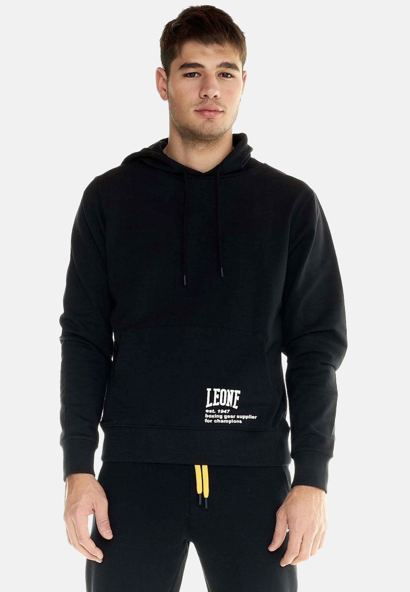 Jeune homme portant un sweat à capuche noir avec une poche avant et le texte blanc "LEONE", debout devant un fond clair uni.