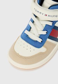 Sneaker montante en suède bleu et matière synthétique beige. Caractéristiques : lacets blancs, bout perforé et rayures rouges et marines sur le côté.
