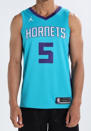Mężczyzna w turkusowej koszulce koszykarskiej Charlotte Hornets z numerem 5 i logo Jordana, stojący z ramionami swobodnie opuszczonymi wzdłuż ciała.