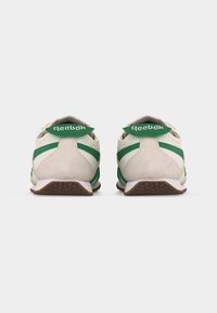 Ζευγάρι μπεζ και πράσινα αθλητικά παπούτσια Reebok, εμφανιζόμενα από πίσω, με έμφαση στις πράσινες ετικέτες στη φτέρνα με λευκά λογότυπα Reebok.