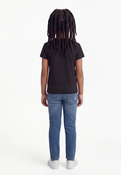 Niño con cabello largo y rizado de espaldas, vistiendo una camiseta negra, jeans azules y zapatillas blancas sobre un fondo blanco.