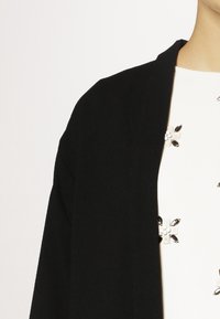 Černý cardigan s jemnou texturou, otevřeným předním designem, s kovovými květinovými akcenty na bílém texturovaném topu pod ním.