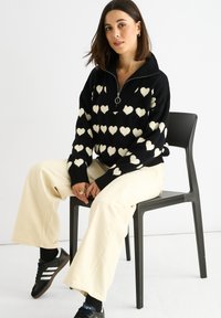 Zwarte trui met wit hartpatroon, kwart-rits ontwerp en ringtrekker; gecombineerd met crèmekleurige wijde broek en zwarte sneakers.