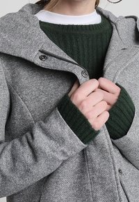 Grijze jas van een wolmix met een rits aan de voorkant, een vast kap en groene geribde gebreide manchetten. Heeft een gestructureerde stof en metalen accenten.