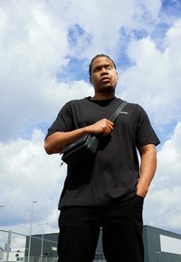 Sort t-shirt og bukser båret af en person, der holder en lille sort crossbody taske. Baggrunden har en skyet himmel og industrielle bygninger.