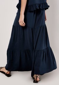 Navy blue tiered maxi skirt with ruffled accents, made from textured fabric, flowing silhouette, and paired with strappy sandals.