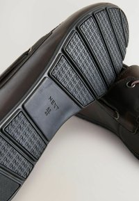 Ruskea nahkakenkien pohja, jossa on kuminen pidempatterni. Ominaisuuksiin kuuluu muotoiltu rakenne ja kohokuvioitu koko- ja mittatieto, mukaan lukien "NEXT" ja mitat.