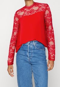 Donna che indossa jeans blu a vita alta e una maglia rossa a maniche lunghe con dettagli in pizzo sulle maniche e sul colletto.