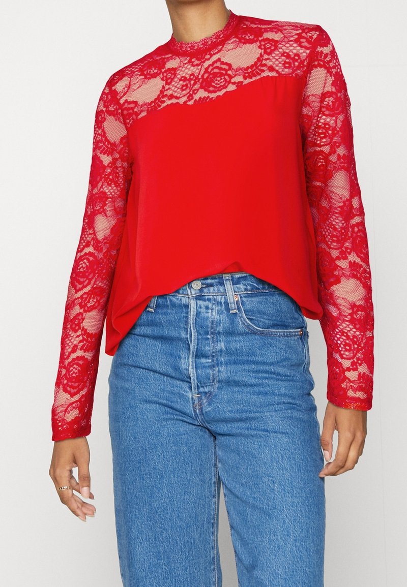 Donna che indossa jeans blu a vita alta e una maglia rossa a maniche lunghe con dettagli in pizzo sulle maniche e sul colletto.