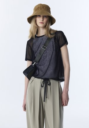 Młoda kobieta w beżowym kapeluszu typu bucket z napisem "WHATEVER", przezroczystej czarnej bluzce na szarym t-shircie, beżowych spodniach i czarnej torebce na pasku na białym tle.