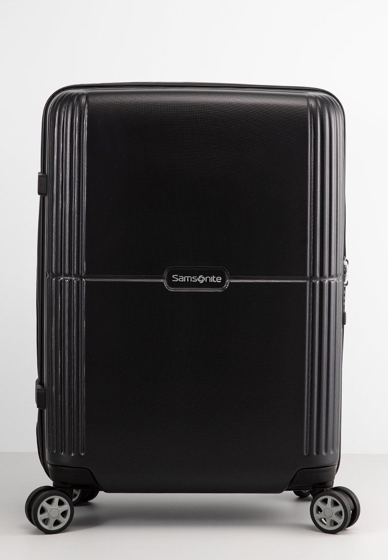 Schwarzer Samsonite-Koffer mit strukturierter Oberfläche, vertikalen Rillen, einem zentralen Logo und vier Rädern für Mobilität. Glatte Oberfläche und rechteckige Form.