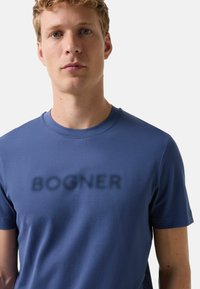 Ung mand med kort blondt hår, iført en blå T-shirt med ordet "BOGNER" trykt i slørede mørke bogstaver hen over brystet.