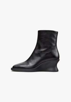 Bota de cuero negro hasta el tobillo con puntera puntiaguda y tacón cuña curvado, mostrada de perfil sobre un fondo blanco.