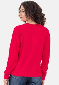 Sweat-shirt rouge à manches longues en tissu doux, avec encolure ronde, épaules tombantes et poignets côtelés. Porté avec un jean bleu.