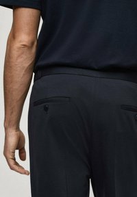 Pantaloni neri con una texture liscia, dotati di due tasche posteriori con chiusura a bottone e una vestibilità sartoriale. La camicia è scura e a maniche corte.
