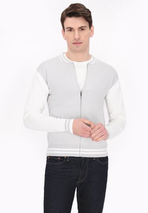 Grijze zip-up trui met witte accenten, geribde manchetten en een sportief ontwerp. Beschikt over een soepele textuur en een getailleerde pasvorm, gedragen over een wit shirt.