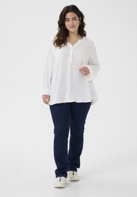 Weißes Langarmhemd mit einem knöpfbaren Ausschnitt und einem strukturierten Muster, kombiniert mit dunkelblauen Jeans und weißen Sneakers.