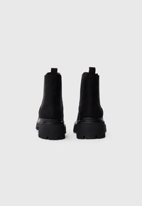 Une paire de bottes Chelsea en suède noir avec de semelles épaisses à crans, vues de l'arrière sur un fond blanc uni.