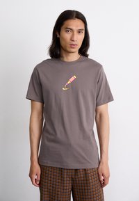 Brun bomulds t-shirt med et farverigt grafik af en flaske og sauce. Designet er enkelt med en rund halsudskæring og korte ærmer.