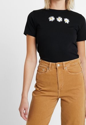 Sort kortærmet t-shirt med tre hvide margueritblomster-applikationer på brystet, parret med højtaljede lysebrune kordfløjlsharem.