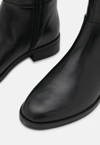 Un par de elegantes botas de tobillo de cuero negro con cremalleras laterales y tacones bajos en un fondo blanco.