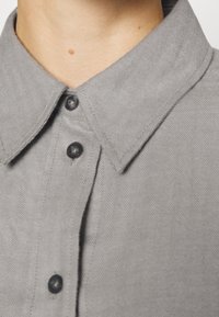 Camicia grigia in misto herringbone con colletto a punta, due bottoni neri e dettaglio in tessuto testurizzato. Vista ravvicinata che mostra il colletto e la posizione dei bottoni.