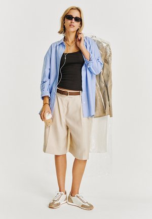 Femme portant des lunettes de soleil, chemise bleue sur un haut noir, short beige, baskets blanches, tenant un café et un sac à vêtements avec des habits protégés par du plastique.