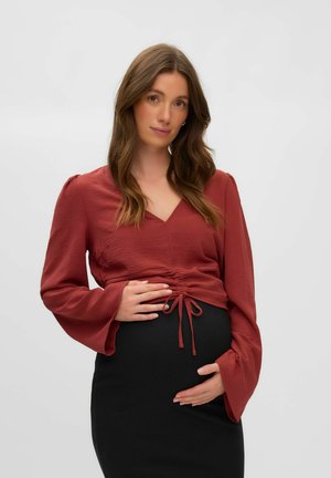 Une femme enceinte portant un chemisier rouge cache-cœur et une jupe noire pose devant un fond blanc uni, les deux mains posées sur son ventre.