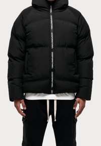 Sort pufferjakke med hætte, frontlynlås med "GOODFORNOTHING" tekst. Vatteret design og en afslappet pasform, båret over en hvid skjorte.