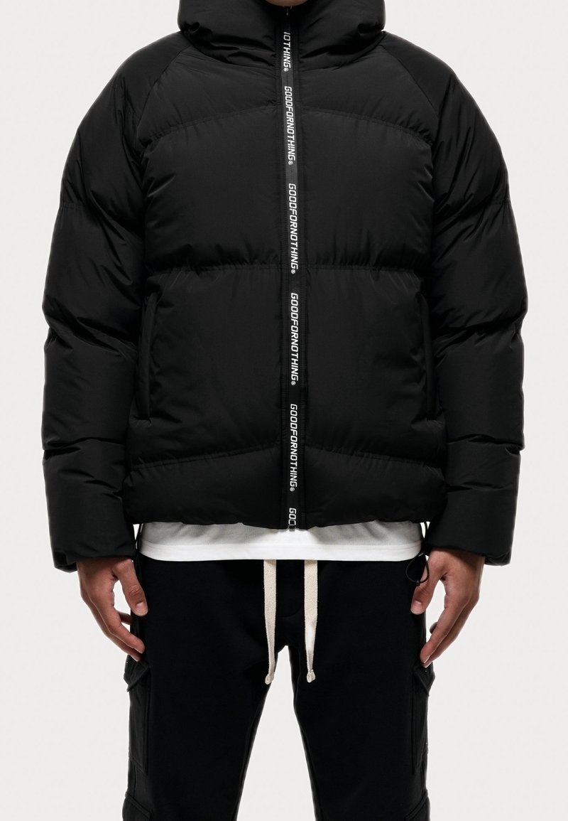 Sort pufferjakke med hætte, frontlynlås med "GOODFORNOTHING" tekst. Vatteret design og en afslappet pasform, båret over en hvid skjorte.