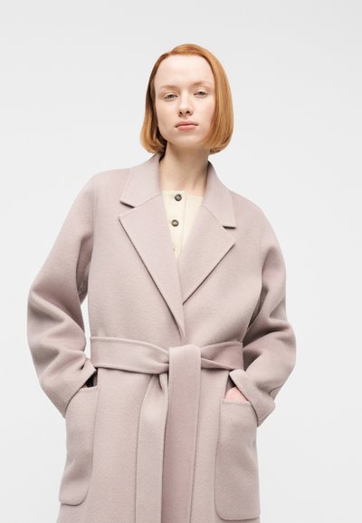 Νεαρή γυναίκα με κοντά κόκκινα μαλλιά, φορώντας ένα ανοιχτό ροζ μάλλινο παλτό με ζώνη, τα χέρια στις τσέπες, με φόντο έναν απλό φωτεινό τοίχο.