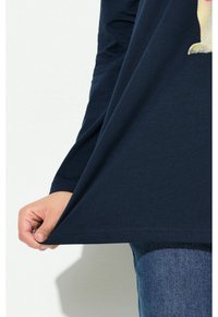 Person, die ein dunkelblaues Langarmshirt mit einem Hundemotiv trägt, das Hemd hält und dehnt, dazu trägt er blaue Jeans.