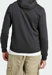 Ciemnoszara bluza z kapturem, wykonana z gładkiego materiału. Ma długie rękawy i dopasowany krój, noszona na białej koszuli i beżowych spodniach typu cargo.
